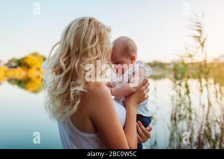 Giorno delle madri. Giovane donna che tiene e bacia la bambina dal fiume primaverile. Famiglia a piedi all'aperto. Bambini che esplorano il mondo Foto Stock