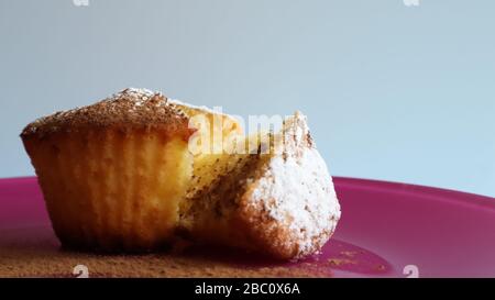 Una cagliata cosparsa di zucchero a velo su un piatto rosa, su sfondo blu, vista laterale. Dessert, un piccolo cupcake. Concetto di cibo: Biscotti al forno bianchi Foto Stock