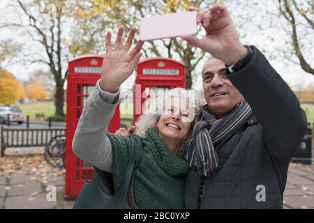 Sorridente coppia senior con selfie nel parco autunnale di fronte alle cabine telefoniche rosse Foto Stock