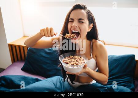 Giovane bella donna nel letto del mattino a casa. La persona affamata divour pasto o cibo. Cereali sani per la colazione con latte nel recipiente. Foto Stock