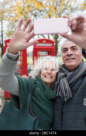 Sorridente coppia senior che prende selfie nel parco di fronte alle cabine telefoniche rosse Foto Stock