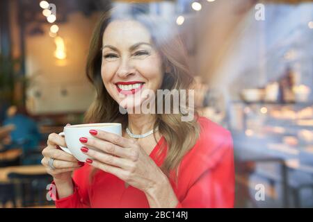 Ritratto di donna matura ridente con tazza di caffè dietro il vetro della finestra Foto Stock