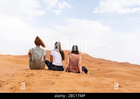 Vista posteriore di tre giovani donne sedute in dune di sabbia nel deserto del Sahara, Merzouga, Marocco Foto Stock