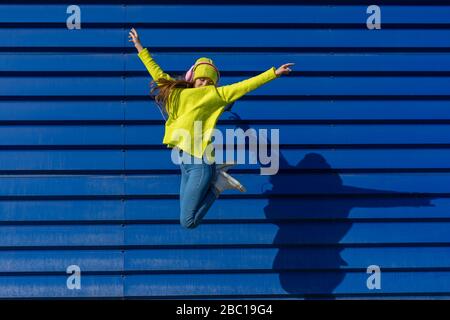 Adolescente ragazza che salta in aria di fronte a sfondo blu ascoltando musica con le cuffie Foto Stock