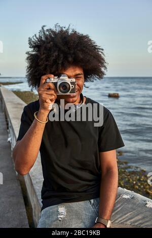 Giovane seduto a muro e fotografando con la sua macchina fotografica sul lungomare Foto Stock