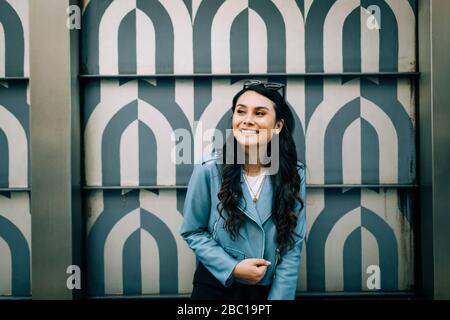 Ritratto di giovane donna con lunghi capelli neri con giacca in pelle blu chiaro davanti alla parete a motivi geometrici Foto Stock