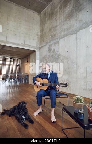 Uomo d'affari senior seduto in ufficio, suonando la chitarra per il suo cane Foto Stock