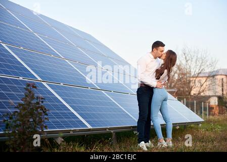 Vista laterale di una bella coppia di bacio in piedi vicino a un grande modulo pv in zona rurale, vista ad angolo basso Foto Stock