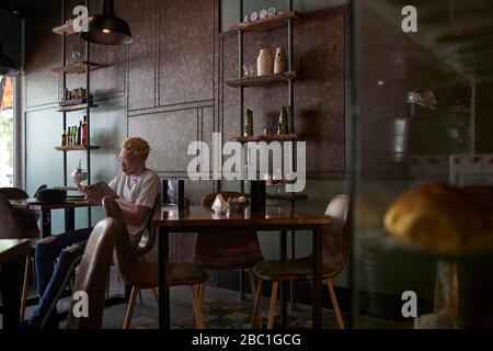 Albino uomo che ha un caffè e che legge un libro in una caffetteria Foto Stock