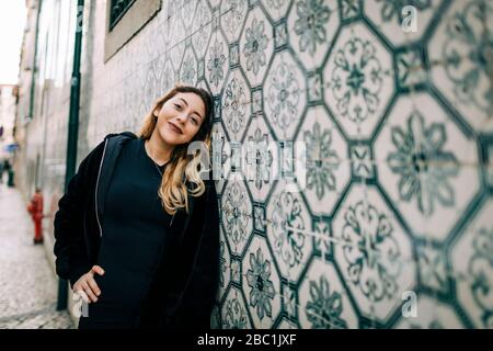 Ritratto di giovane donna felice vestito di nero appoggiato contro il muro di Azulejo, Lisbona, Portogallo Foto Stock