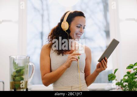 Adatta donna in piedi in cucina, preparando frullato sano, usando la ricetta in linea Foto Stock