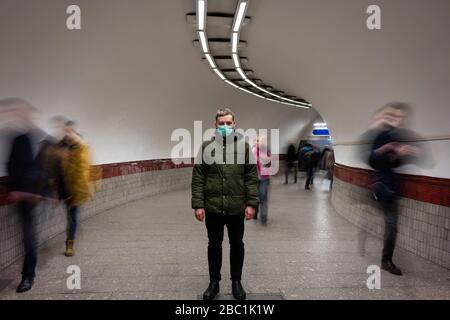 Giovane uomo con maschera facciale in piedi isolato nel sottopassaggio della metropolitana, con persone che si muovono intorno a lui Foto Stock