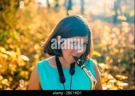 allegra giovane donna con capelli scuri in abiti blu suona il sassofono in autunno parco giallo Foto Stock