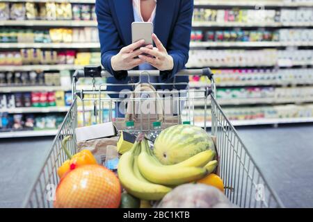 Donna con smart phone che spinge il carrello in supermercato Foto Stock