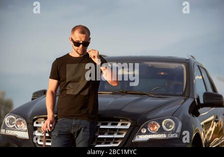 Ritratto di un uomo sicuro di sé di successo in piedi di fronte alla sua bella auto nera, indossando occhiali da sole scuri, maglietta nera e jeans, primo piano Foto Stock