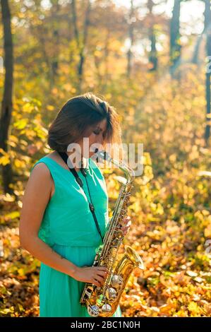 cute ragazza dai capelli neri in un abito blu gioca il sassofono giallo nella foresta autunnale Foto Stock