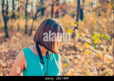 lateralmente da vicino una donna bruna in un abito blu gioca il sassofono in un parco autunnale giallo Foto Stock