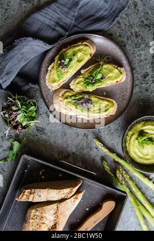 Fette di pane con avocado vegetariano, zucchine e asparagi si spargero, sormontate da piselli e germogli di ravanello su un piatto di ceramica Foto Stock