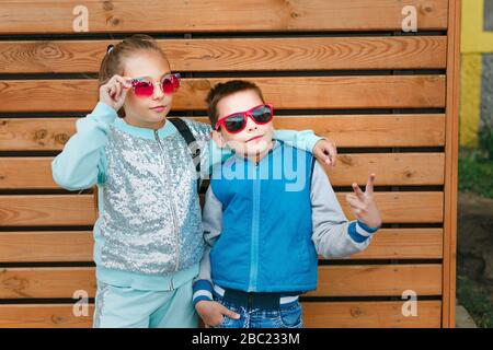 Fashion kids in città sorge su una parete in legno. Alla moda di un ragazzo e una ragazza in occhiali da sole permanente sulla strada Foto Stock