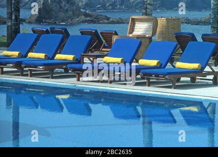 Fila di sedie a sdraio blu con asciugamani gialli allineati lungo il lato di una piscina in un hotel a Careyes, Jalisco, Messico. Essi sono riflessi nell'acqua. Foto Stock