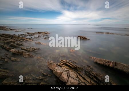 Un tranquillo mare a lunga esposizione di nuvole e onde in movimento, che crea una bella nebbia di alce intorno alle suggestive rocce in primo piano, prese in po Foto Stock