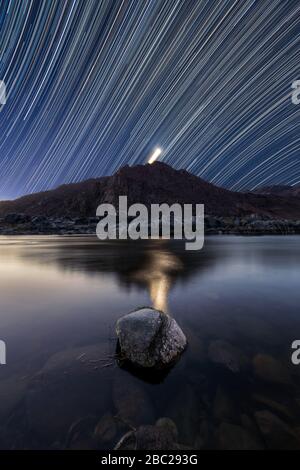 Un bellissimo paesaggio verticale notturno con sentieri stellari contro un cielo blu profondo e la luna dietro le montagne, che si riflette nella Rive arancione Foto Stock