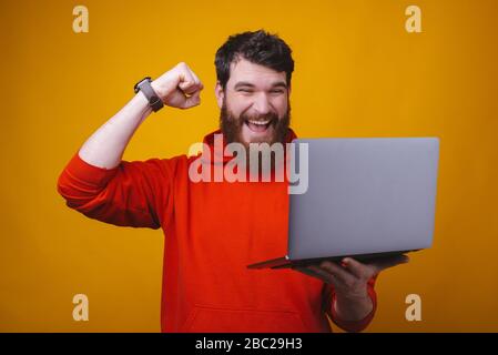 Ho vinto. La foto di un giovane uomo bearded sta mostrando il gesto del vincitore mentre guarda il suo laptop che tiene. Foto Stock