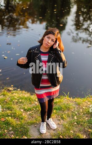 bella ragazza adolescente in sneakers bianche, un abito a maglia a righe rosse e una giacca nera, raddrizza i capelli scuri, in piedi accanto al fiume Foto Stock