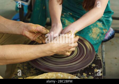 Mani abbronzate maschili del padrone e le mani di una ragazza in un grembiule verde su una ruota di Potters fanno una brocca di argilla gialla Foto Stock