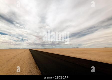 Uno spaventoso paesaggio desertico preso vicino a Luderitz, Namibia, con la strada nazionale del catrame nero che conduce alla distanza, sabbia del deserto in primo piano e. Foto Stock