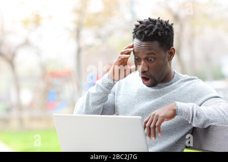 L'uomo nero stupito che legge le notizie sorprendenti sul laptop seduto al banco del parco Foto Stock