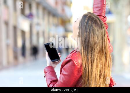 Vista posteriore di una ragazza eccitata che tiene il telefono e mostra uno schermo vuoto nella strada Foto Stock
