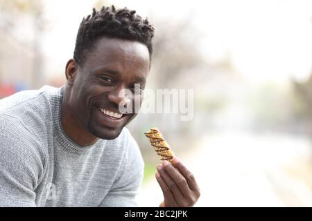 Uomo nero felice che tiene granola bar pronto per mangiare guardando la macchina fotografica in un parco con spazio copia Foto Stock