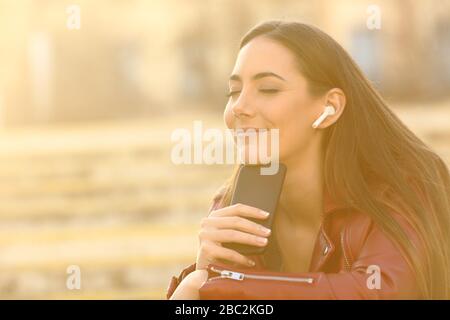 Donna rilassata che indossa gli auricolari che ascoltano la musica sullo smartphone con occhi chiusi e una luce calda Foto Stock