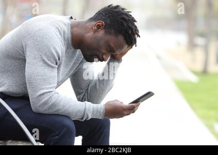 Vista laterale ritratto di un triste uomo nero lamentando il controllo del telefono cellulare seduto su una panchina in un parco