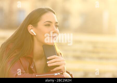Donna rilassata che indossa gli auricolari ascolta la musica sul telefono cellulare guardando lontano in strada con una luce calda del sole Foto Stock