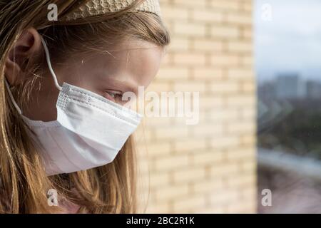 Coronovirus Quarantine Concept.A ragazza in una maschera protettiva dal virus sulla sua faccia siede vicino alla finestra, Coronavirus .Sad child.Stay a casa Foto Stock