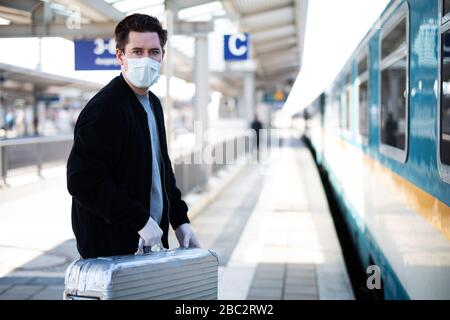 Uomo con una valigia sulla piattaforma, ha una maschera facciale e guanti contro l'infezione con il virus corona Foto Stock