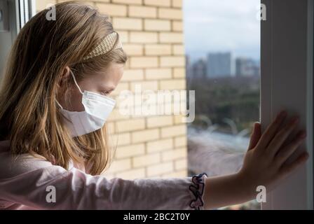 Una ragazza in una maschera protettiva dal virus sul viso si siede vicino alla finestra, guardando su Street.Prevention Epidemic.Sad bambino ragazza in maschera medica dal Foto Stock