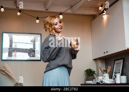 Sorridente giovane donna in abbigliamento casual in piedi in cucina, tenendo mestolo e frusta Foto Stock
