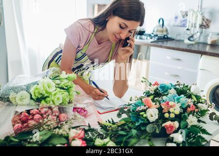 Lavoro da casa durante la quarantena del coronavirus. Donna fiorista compone bouquet di fiori riceve ordini al telefono. Consegna Foto Stock