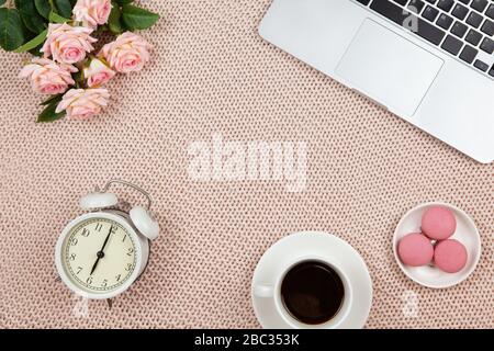 Telelavoro da casa. Moderno spazio di lavoro femminile, vista dall'alto. Laptop, caffè, torte, rose, orologio su coperta a maglia, spazio copia, piatto lay. Scrivania Foto Stock