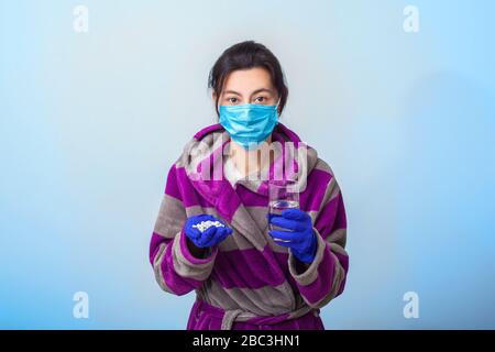 Una ragazza in una maschera medica protettiva e guanti, tiene tra i suoi palmi una manciata di pillole e un bicchiere d'acqua. Sfondo azzurro. In una maschera respiratoria. Il concetto di coronavirus e covid-19 Foto Stock