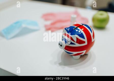 Una banca piggy con una bandiera inglese, guanti, maschera, una mela e un igienizzatore per le mani sul tavolo. COVID - 19 prevenzione Foto Stock