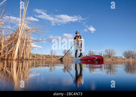 Il rematore maschile senior in una muta sta remando una pagaia in piedi su su su un lago in Colorado, paesaggio di inverno o di primavera presto, telecamera d'azione di angolo basso vie Foto Stock