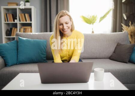 Vista frontale di una donna matura in un maglione giallo alla moda seduto sul divano e al lavoro sul computer portatile. Donna sorridente con capelli biondi usando il computer per remo Foto Stock