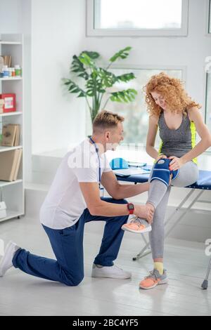 Fisioterapista maschile inginocchiato su un ginocchio, esaminando la gamba della paziente femminile Foto Stock