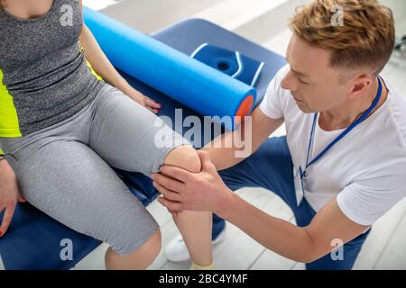 Fisioterapista maschile che cura le lesioni del ginocchio del paziente femminile seduto sul divano Foto Stock