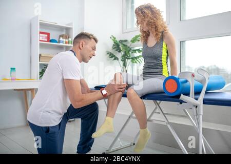 Fisioterapista maschile inginocchiato su un ginocchio, esaminando il ginocchio doloroso della paziente femminile Foto Stock