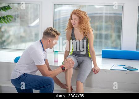 Fisioterapista maschile inginocchiato su un ginocchio, trattamento doloroso ginocchio di paziente femminile seduta su davanzale Foto Stock